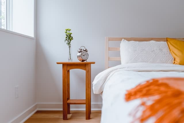 Half of a made bed with a wooden night stand on the left with a plant and alarm clock on top